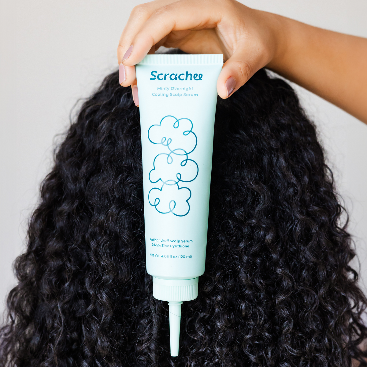 A person's hand holding a tube of 'Scrachee' hair serum with a blue label, above a background of curly hair.