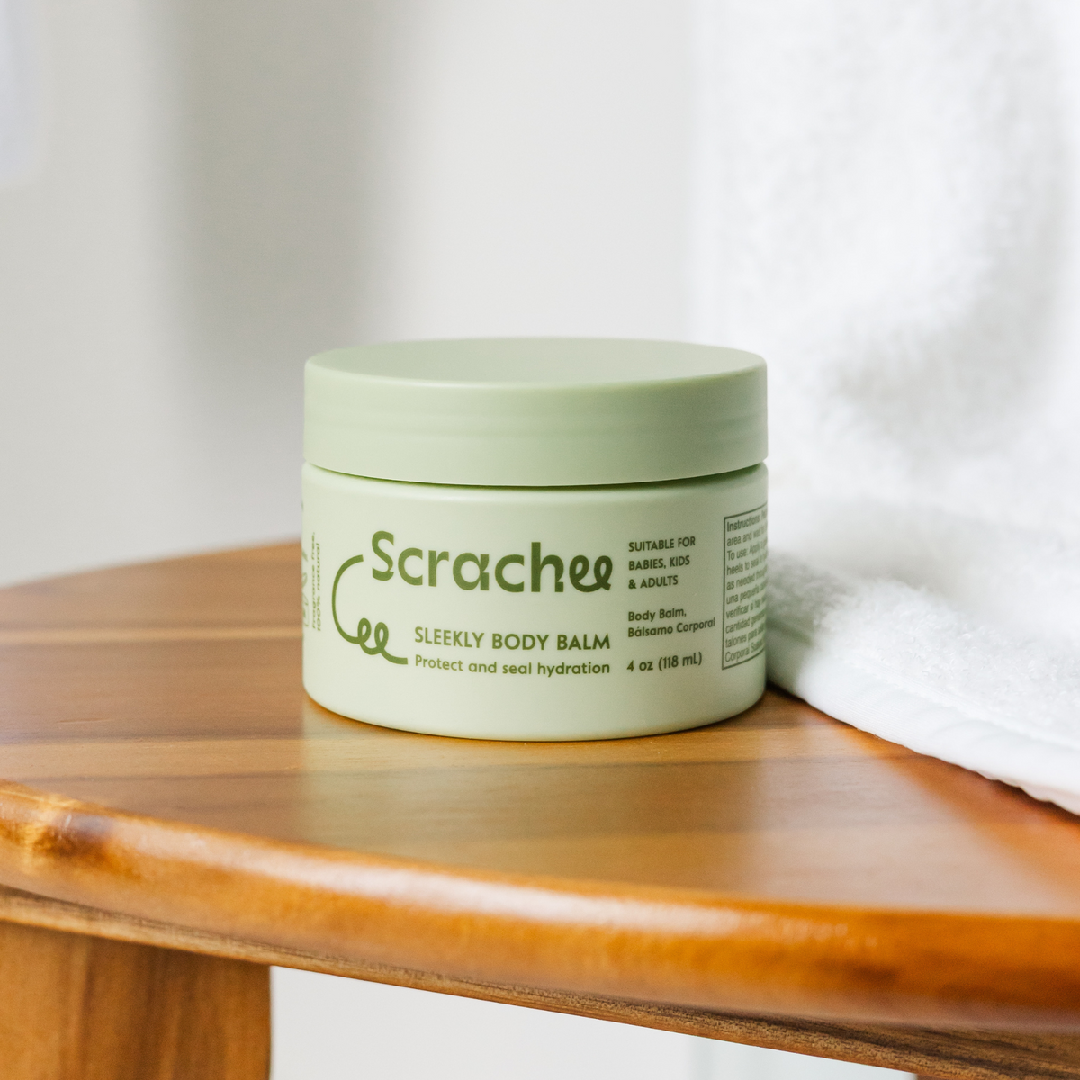 Green jar of Scrachee Sleekly Body Balm on a wooden surface with a white towel in the background.
