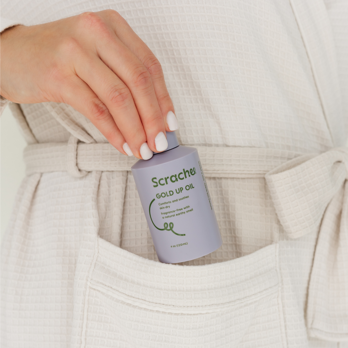 Hand holding a bottle of 'Scrachee' Gold Up Oil with a neutral background