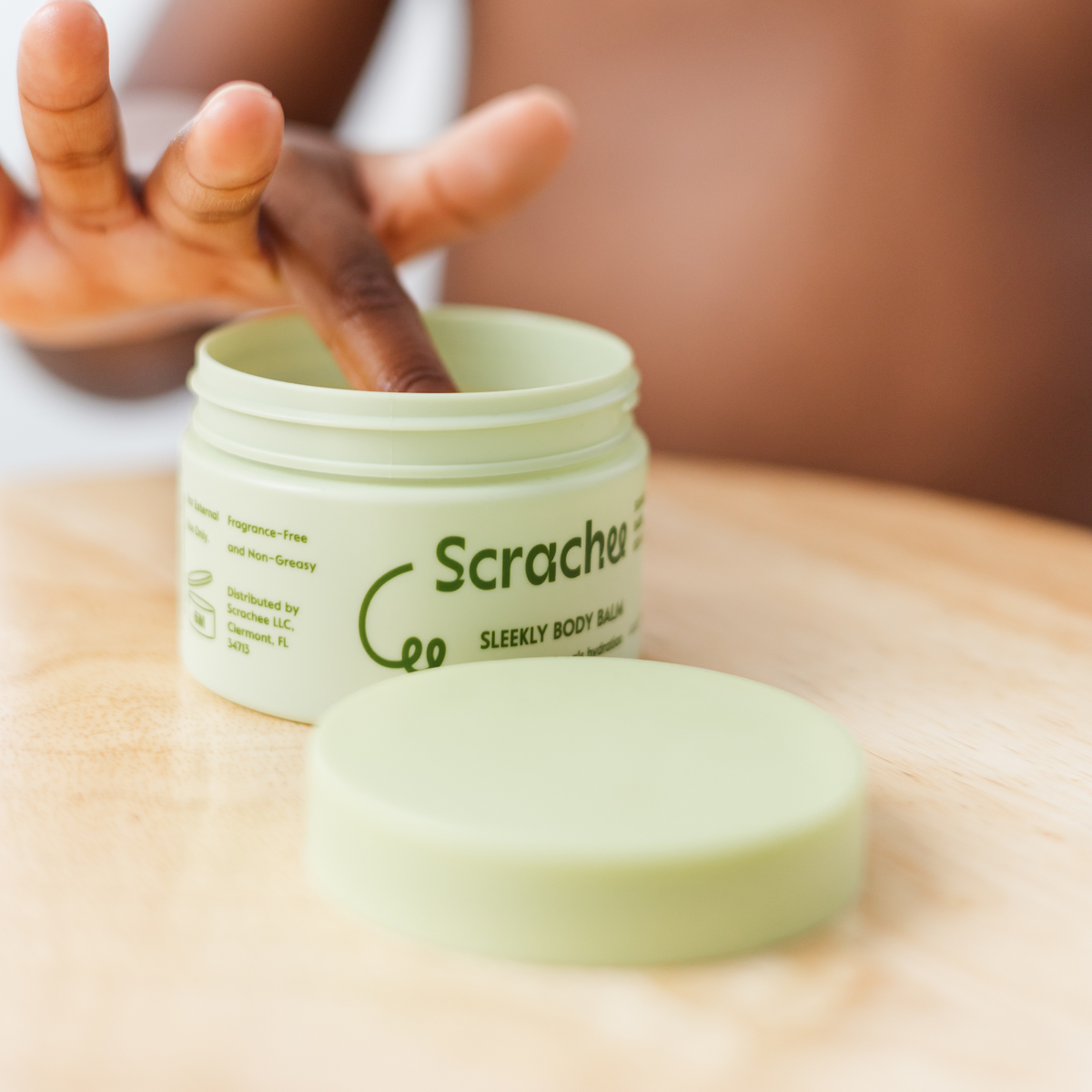 Green jar of 'Scrachee' body balm on a wooden surface with a boy dipping his hands on it.